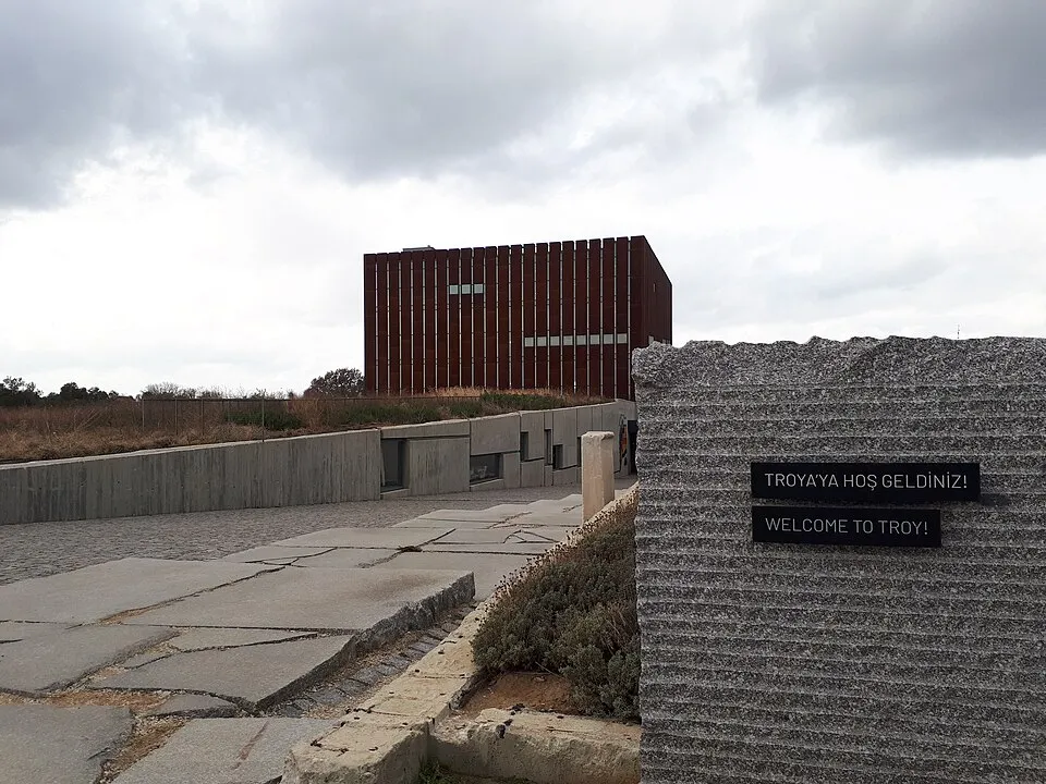 Troya Müzesi'nin konumunu belirten bu görselde, bulutlu bir gökyüzünün altında, pas rengi modern müze binasına doğru ilerleyen düzensiz taş plakalarla döşeli giriş yolu ve yolun sağında yer alan, üzerinde Türkçe ve İngilizce 'TROYA'YA HOŞ GELDİNİZ!' yazıları bulunan gri, dokulu bir karşılama duvarı öne çıkıyor.
