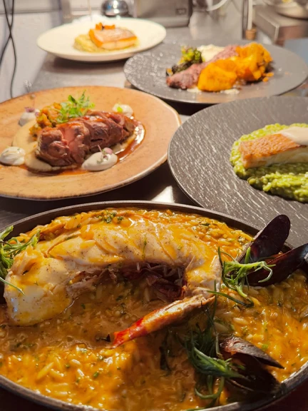 Patmos'taki Vangelis restoranında sunulan, ortada büyük bir balık yemeğiyle birlikte çeşitli et ve deniz mahsulleri seçeneklerini gösteren bir masa düzenlemesi: ön planda arpa şehriyesi üzerinde servis edilen bütün bir balık, midyeler ve karideslerle zenginleştirilmiş, arka planda ise et yemekleri ve balık fileto içeren diğer gurme sunumlar yer alıyor.