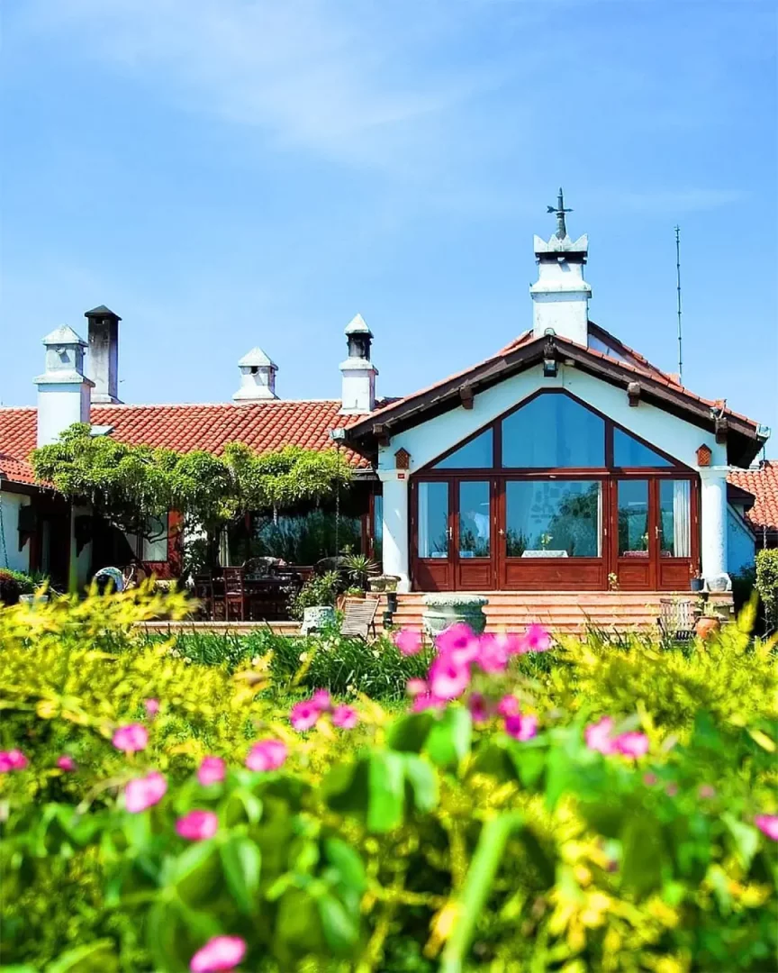 Çatalca'daki bu villa, ön planda yemyeşil bahçesindeki pembe çiçeklerle, kiremit çatılı, beyaz duvarlı, geniş cam cepheli ve birkaç bacalı yapısıyla güneşli mavi gökyüzü altında dingin bir manzara sunuyor.