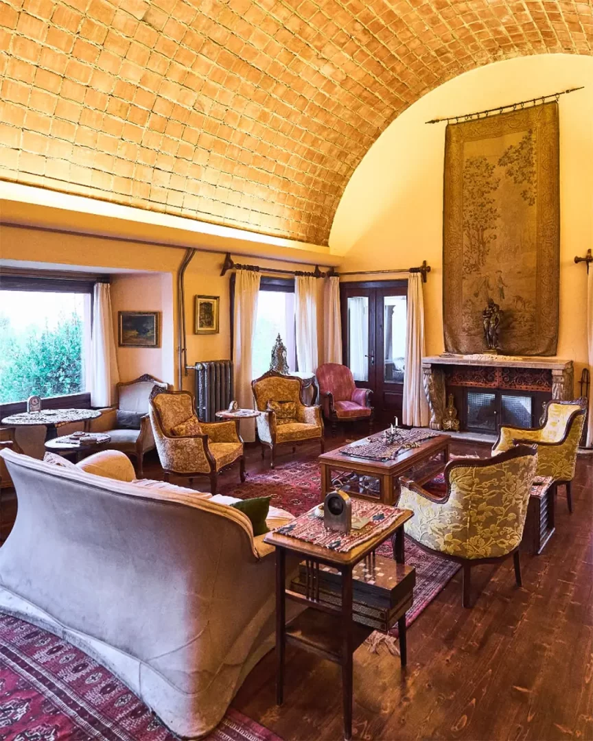 Kemerli tuğla tavanlı ve ahşap zeminli geniş bir villa salonunda, bej kanepe, desenli berjerler, ahşap sehpalar ve şöminenin üzerindeki büyük goblen duvar halısıyla döşenmiş sıcak ve davetkar bir oturma odası manzarası.