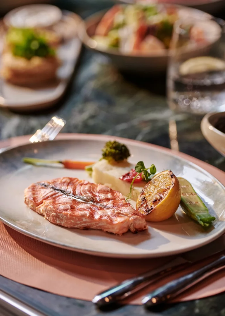 Beyaz bir tabağın ortasında yer alan, ızgara izleri belirgin pembe renkli bir somon fileto, yanında kremsi patates püresi, yeşil brokoli, kızarmış havuç, ızgara limon ve kabak dilimleriyle özenle hazırlanmış bu deniz ürünleri tabağı, Voyage Kundu Blue Restoran'ın özel lezzetlerinden birini sunuyor.