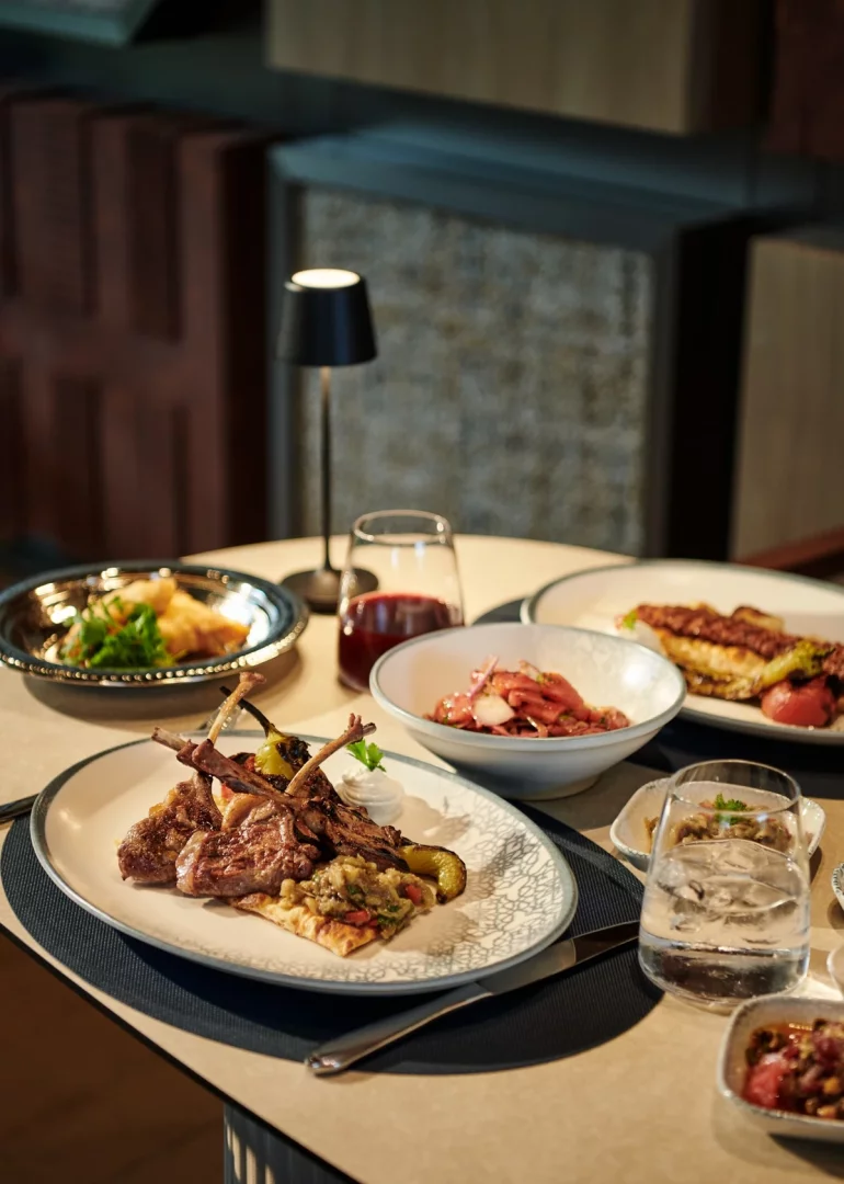Voyage Kundu Kebappa Restoran'da akşam yemeği menüsünden seçilen, kemikli kuzu pirzola, közlenmiş biber, patlıcan ezmesi ve çeşitli mezelerle hazırlanan ana yemek tabağı ile kırmızı şarap ve su bardaklarının bulunduğu, loş ışıklı şık bir masa.