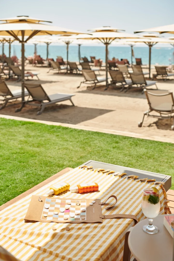 Resort plaj deneyimini yansıtan bu karede, sarı beyaz çizgili bir havlu üzerinde güneş kremleri, bir oyun tahtası ve yanında serinletici bir kokteyl masada dururken, arka planda uzanan kumsalda çok sayıda şezlong ve şemsiye denize nazır uzanmaktadır.
