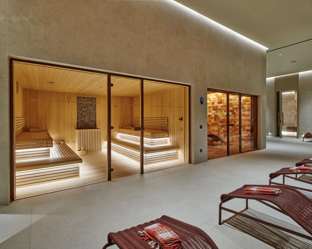 Modern ve aydınlık bir spa ortamında, içindeki basamaklı ahşap oturma alanları ve zarif aydınlatmalarıyla dikkat çeken cam kapılı sauna ile yanındaki tuz duvarlı oda, Voyage Kundu Sense Spa'nın huzurlu ambiyansını yansıtan havlu serili şezlonglarla bir arada görülüyor.