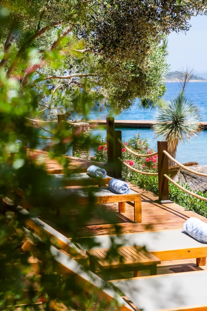 WAM Hotel Bodrum'un Demirbükü'ndeki doğal güzelliklerle çevrili ahşap iskele ve deniz kenarı dinlenme alanında, katlanmış havluların yer aldığı konforlu şezlonglar, otelin lüks yerine kâfi bir Akdenizli yaşam anlayışını yansıttığı huzurlu bir anı sunuyor.