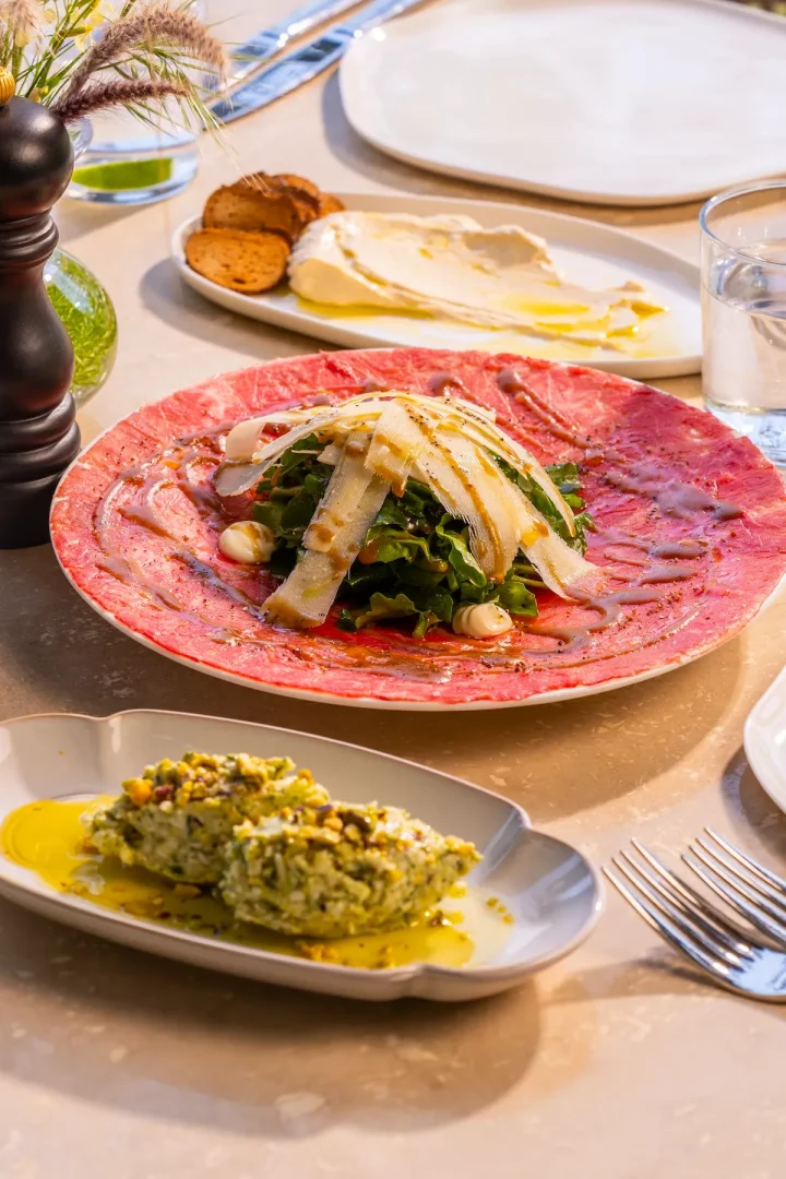 Masada sunulan iştah açıcı bir sunumda, ana odak noktası olan geniş kırmızı tabakta roka, ince dilimlenmiş parmesan peyniri ve sos ile taçlandırılmış taze carpaccio di manzo yer alırken, ön planda zeytinyağı ve fıstıklarla süslenmiş yeşil bir meze ve arka planda hafif bir humus ile çıtır ekmek dilimleri tamamlayıcı bir görünüm sunuyor.