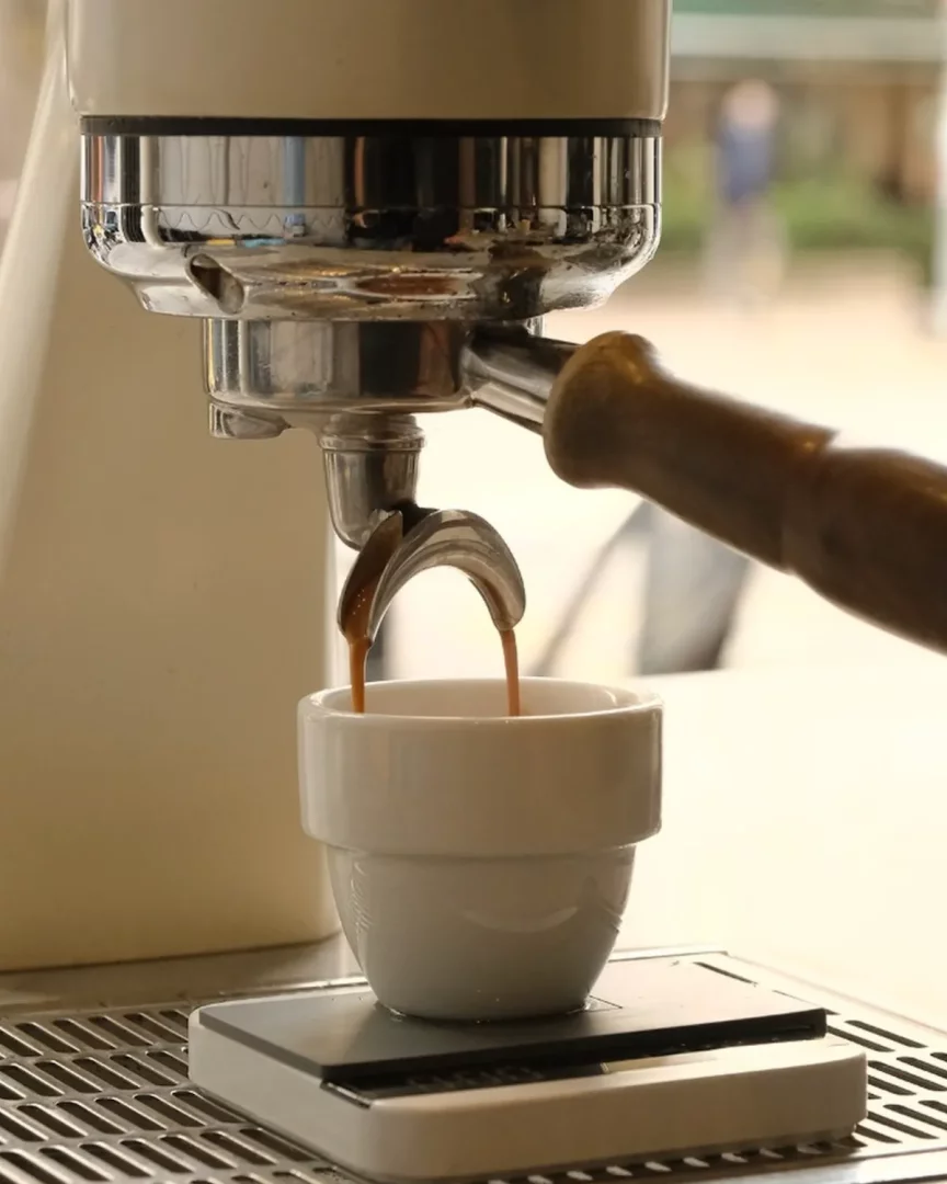 Bir espresso makinesinden portafiltrenin çift oluklu çıkışından küçük, şık beyaz bir fincana yavaşça akan koyu kahve, en iyi kahvecilerde deneyimleyebileceğiniz kaliteli kahve hazırlama sürecini canlı bir şekilde gösteriyor.