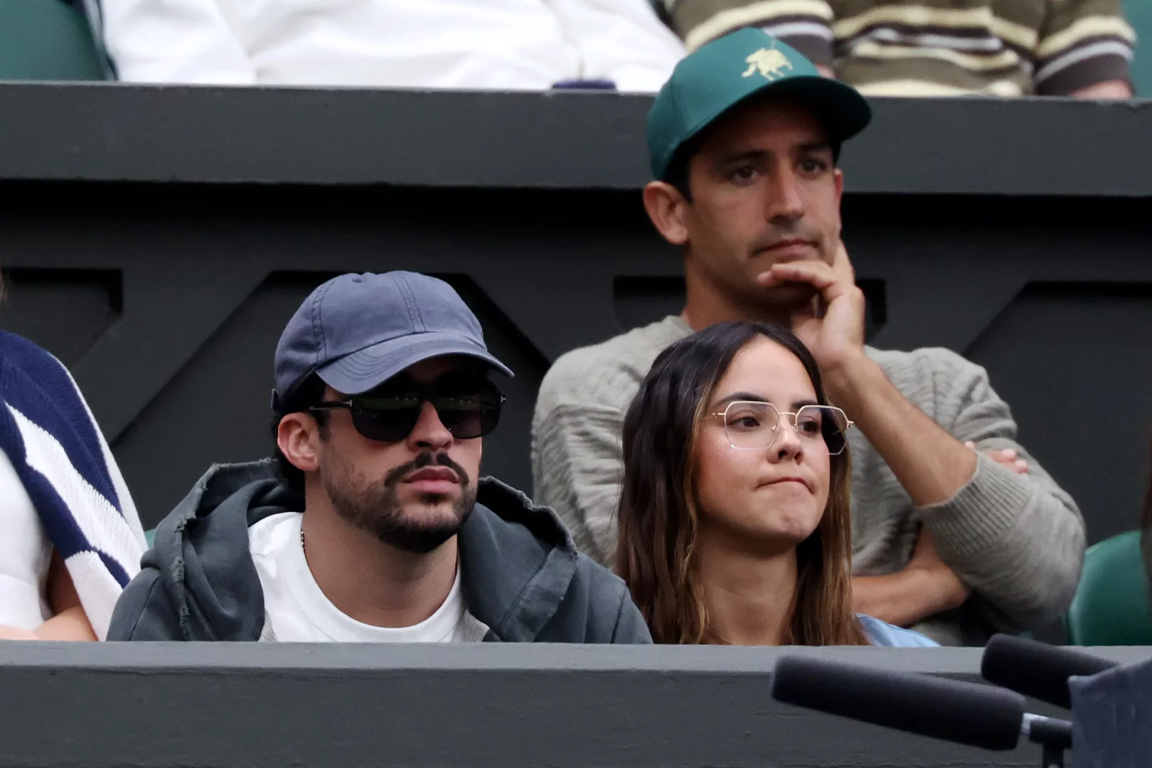 Wimbledon tenis turnuvasının prestijli tribünlerinde, koyu şapkalı ve güneş gözlüklü Bad Bunny ile yanında gözlüklü bir kadın ve arkada başka bir erkek, İngiliz yazının bu ikonik ritüelini dikkatle takip ederken, zengin atmosferin bir parçası oluyorlar.