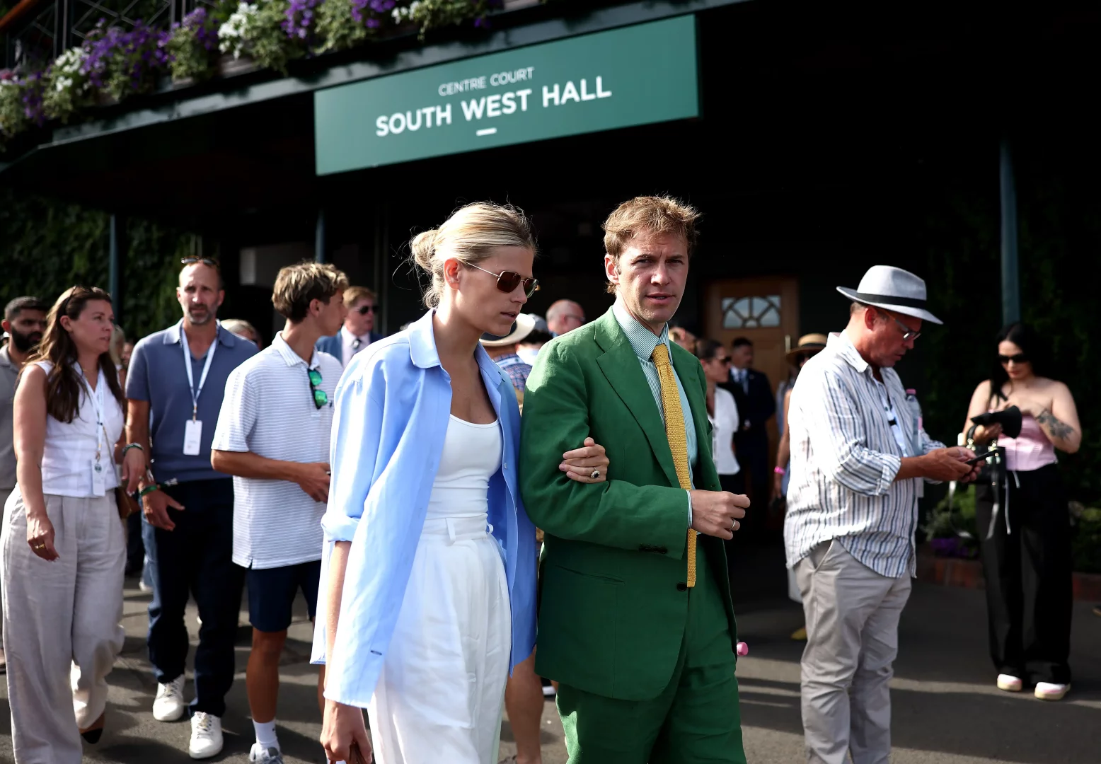 Wimbledon'ın ikonik Centre Court Güney Batı Salonu tabelası altında, zarif stiliyle dikkat çeken, açık mavi gömlekli ve beyaz pantolonlu sarışın bir kadınla, yemyeşil takım elbiseli bir erkeğin kol kola yürüdüğü an, İngiliz yazının prestijli tenis turnuvası atmosferini yansıtan kalabalık bir sahneyi betimliyor.