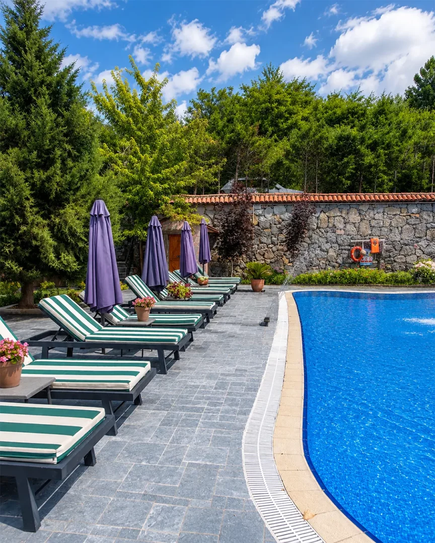 Wineport Lodge Ağva Şile'deki açık havuz alanında, parlak mavi suyun kıvrımlı kenarları boyunca dizilmiş yeşil-beyaz çizgili minderli şezlonglar ve mor şemsiyeler, ağaçlarla çevrili taş bir duvarın önünde, güneşli bir günde huzurlu bir dinlenme ortamı sunuyor.