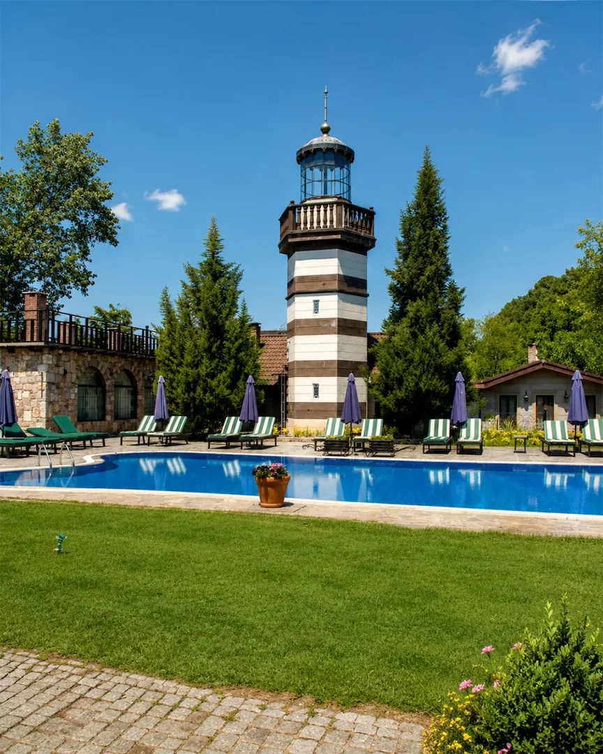Ağva'da bulunan Wineport Lodge'un mimarisini yansıtan kahverengi-beyaz çizgili deniz feneri görünümlü kulesinin önünde, masmavi bir yüzme havuzu etrafında sıralanmış şezlonglar ve lacivert şemsiyelerle donatılmış yemyeşil bahçe, huzurlu bir tatil ambiyansı sunuyor.