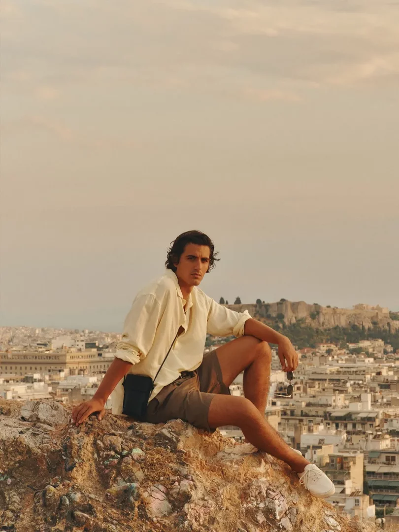 Şehir manzaralı kayalık bir tepede, gün batımı ışığında oturan, krem rengi gömlek ve şort giymiş genç model, arkasında Akropolis silüetiyle, ayağındaki beyaz bağcıklı kanvas ayakkabı modeli ile hem rahat hem de şık bir stil sergiliyor.