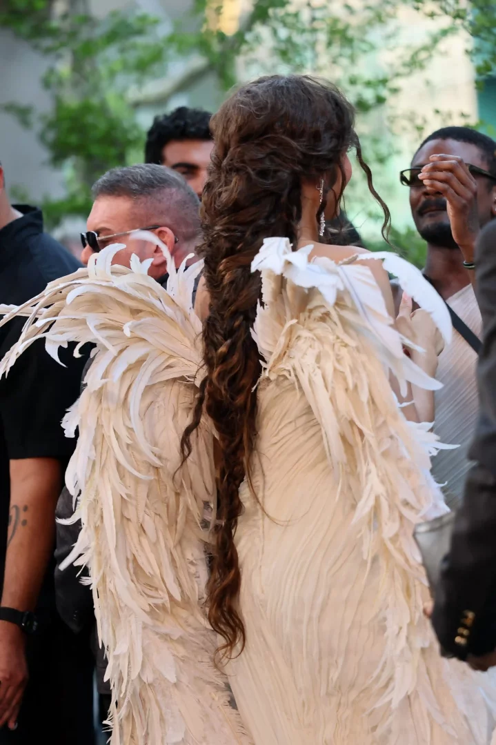 Zendaya, The Odyssey New York prömiyerinde sırtı dönük olarak görülüyor; uzun, dalgalı örgülü saçları ve omuzlarından sırtına doğru uzanan, zarif krem rengi tüy kanat detaylarıyla süslü elbisesi kalabalığın arasında dikkat çekiyor.