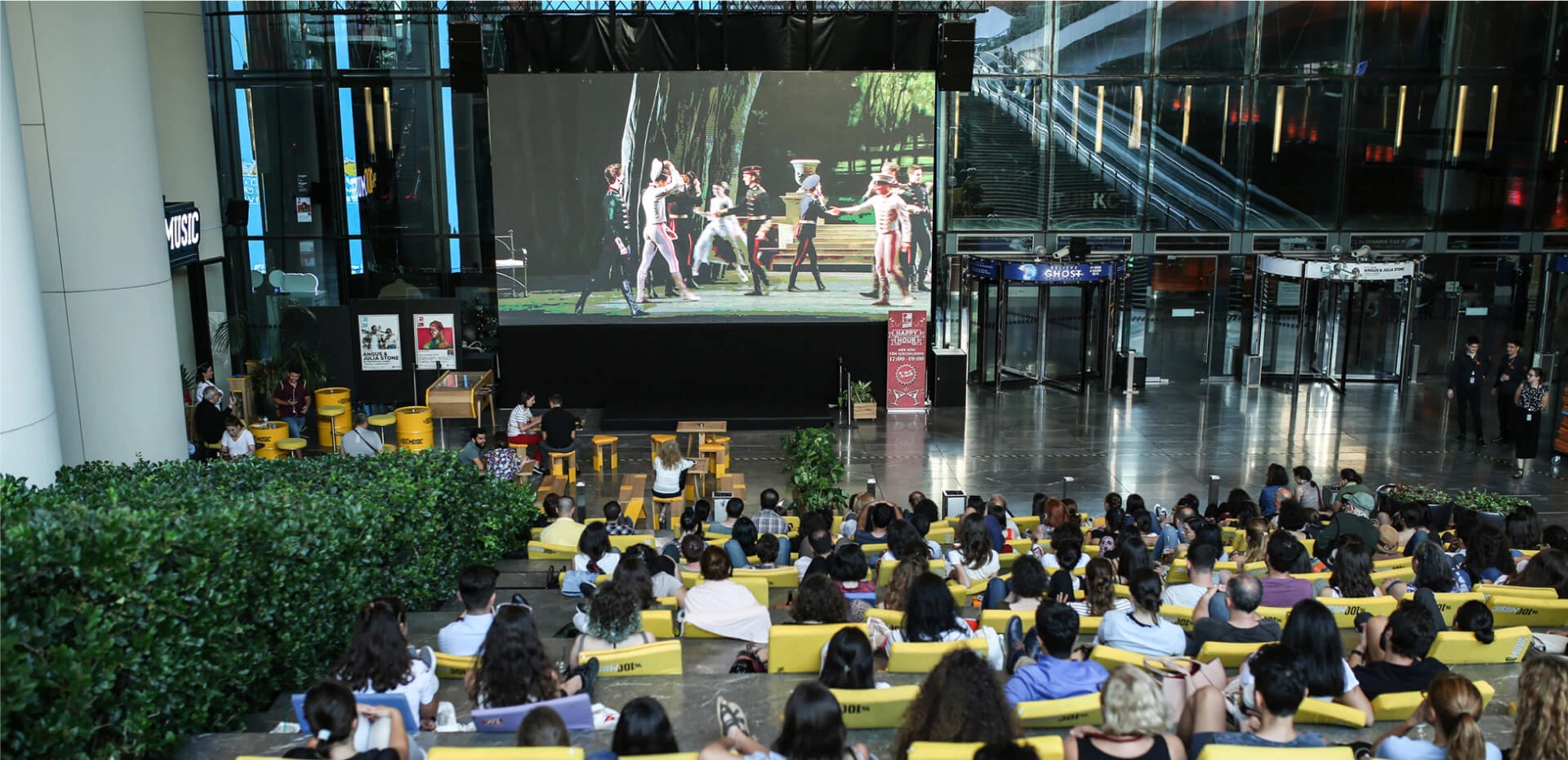 Zorlu PSM'de düzenlenen açık hava sinema etkinliğinde, geniş bir ekranda bale benzeri bir sahne gösterisi izleyen kalabalık izleyici topluluğu, sarı minderler ve tabureler üzerinde rahatça otururken, arka planda modern bir binanın cam duvarları ve yürüyen merdivenleri yansıyor.
