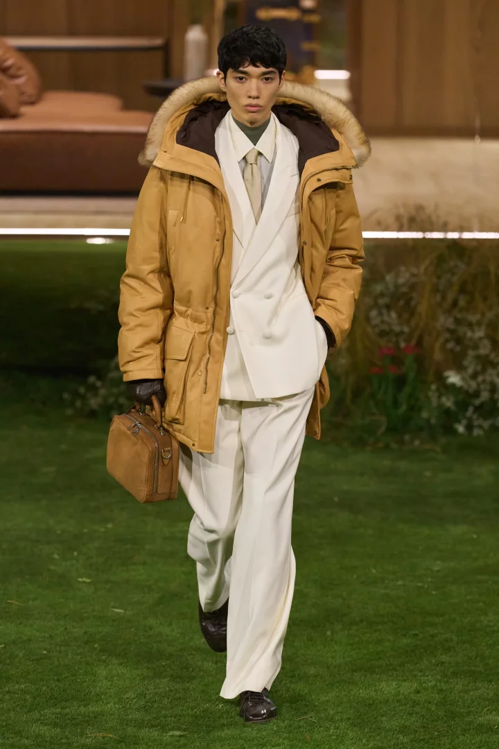 Louis Vuitton'un 2026 sonbahar erkek modası koleksiyonundan bir model, podyumda devetüyü rengi kürklü parkası, krem takım elbisesi, zeytin yeşili gömleği ve kravatıyla, elinde küçük kahverengi bir evrak çantasıyla yürüyor.