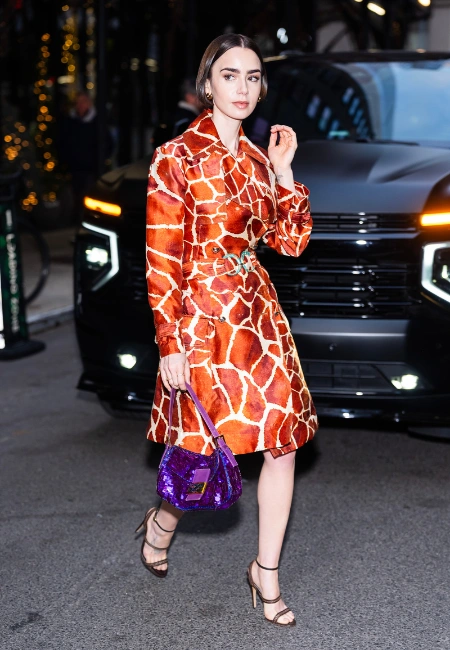 Lily Collins, turuncu-kahverengi ve beyaz zürafa desenli, kemerli, şık bir palto elbise içinde, göz alıcı mor pullu ikonik Fendi Baguette çantasını sol elinde tutarak, ince bantlı topuklu sandaletleriyle kaldırımda zarifçe yürürken objektife bakıyor.