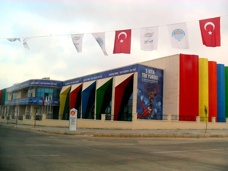 Mersin'de düzenlenen 2013 Akdeniz Oyunları'na ev sahipliği yapan tesislerden biri olduğu anlaşılan, ön cephesinde Türk bayrakları ve 3 Kıta Tek Yürek sloganlı dev afişler asılı, gökkuşağı renklerinde panellerle süslü modern bir binanın dışarıdan çekilmiş geniş açılı görüntüsü.