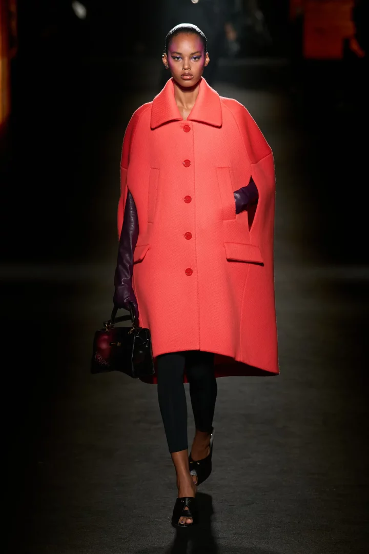 Model, 2026 Balenciaga Sonbahar-Kış defilesinde parlak mercan rengi pelerin palto, uzun mor eldivenler, siyah pantolon ve topuklu ayakkabıları ile elinde siyah bir çanta taşıyarak, bu sezonun öne çıkan renk trendlerini göz alıcı bir şekilde sunuyor.