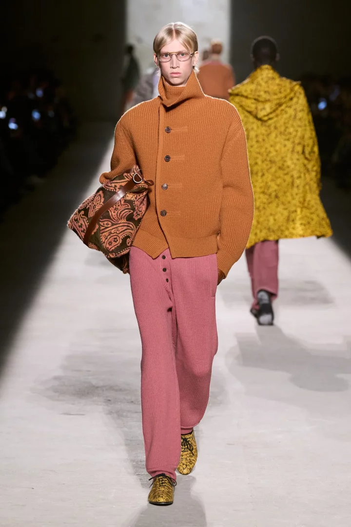 Podyumda yürüyen bir erkek model, 2026 sonbahar renk trendlerini yansıtan alev turuncusu renkli örgü hırkası, toz pembe çizgili pantolonu, paisley desenli kahverengi çantası ve sarı desenli ayakkabılarıyla erkek modasında dikkat çekici bir stil sergiliyor.