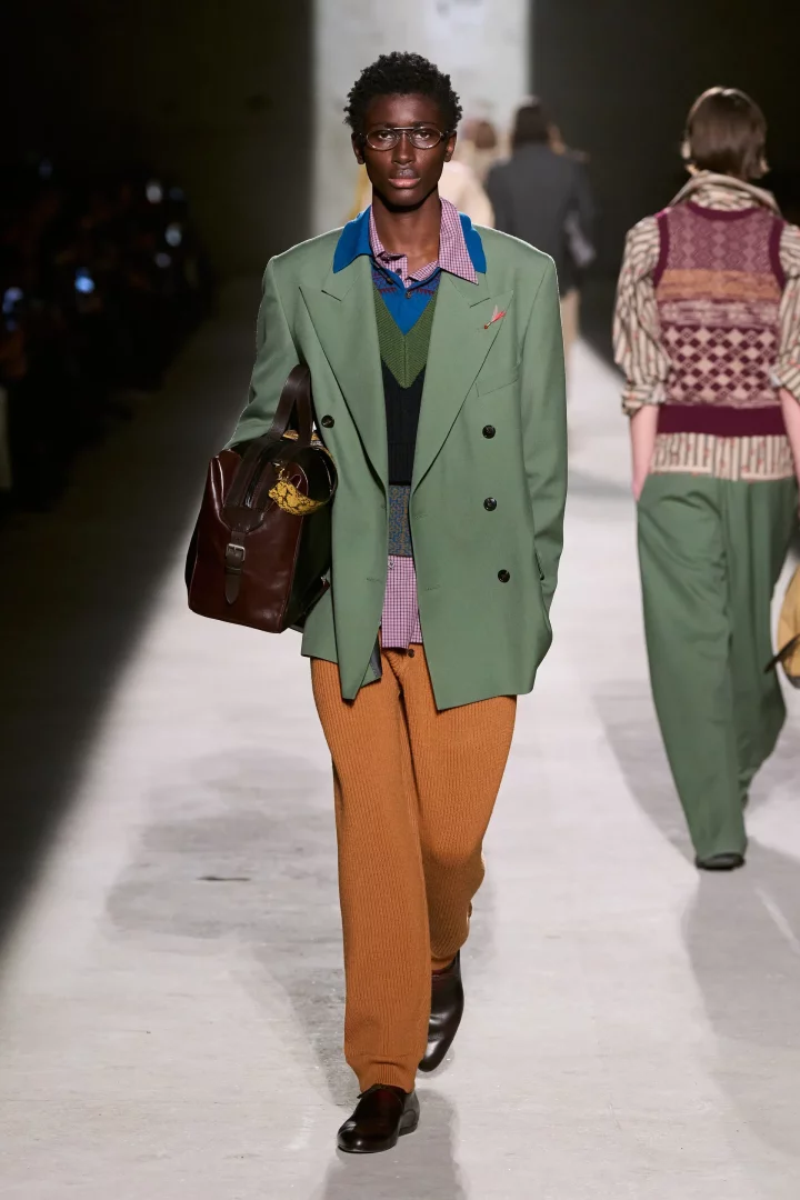 Podyumda yürüyen bir erkek model, 2026 sonbahar renk trendlerini yansıtan, zeytin yeşili blazer, mavi-yeşil süveter yelek ve alev turuncusu pantolondan oluşan katmanlı, şık bir erkek modası kombiniyle kahverengi bir çanta taşıyor.