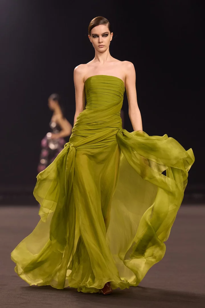 Elie Saab defilesinden, 2026 sonbahar renk trendleri arasında öne çıkan şartröz renginde, uçuşan kumaşlardan yapılmış straplez bir elbiseyle koyu podyumda yürüyen modelin zarif duruşu, elbisenin hareketli ve hafif dokusunu vurguluyor.