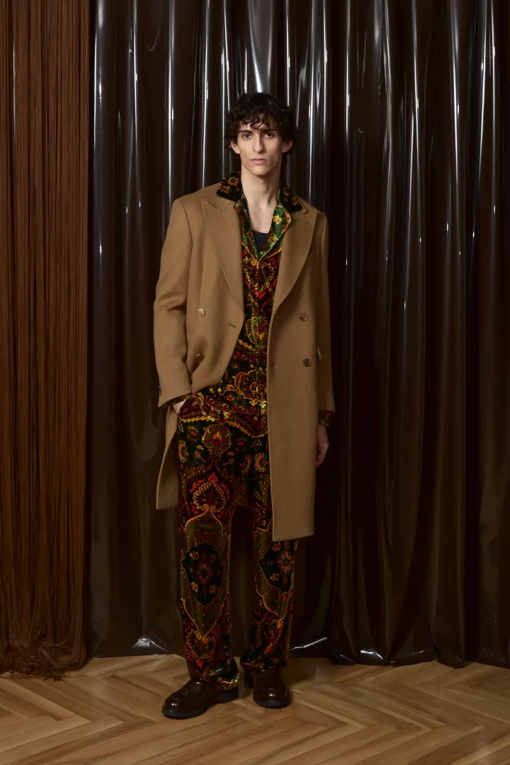 Etro markasının 2026 sonbahar erkek renk trendleri koleksiyonundan, kahverengi perdelerin önünde duran bir model; devetüyü rengi kruvaze paltosu ve canlı kırmızı, turuncu, yeşil tonlarında zengin etnik desenli takımıyla dikkat çekiyor.