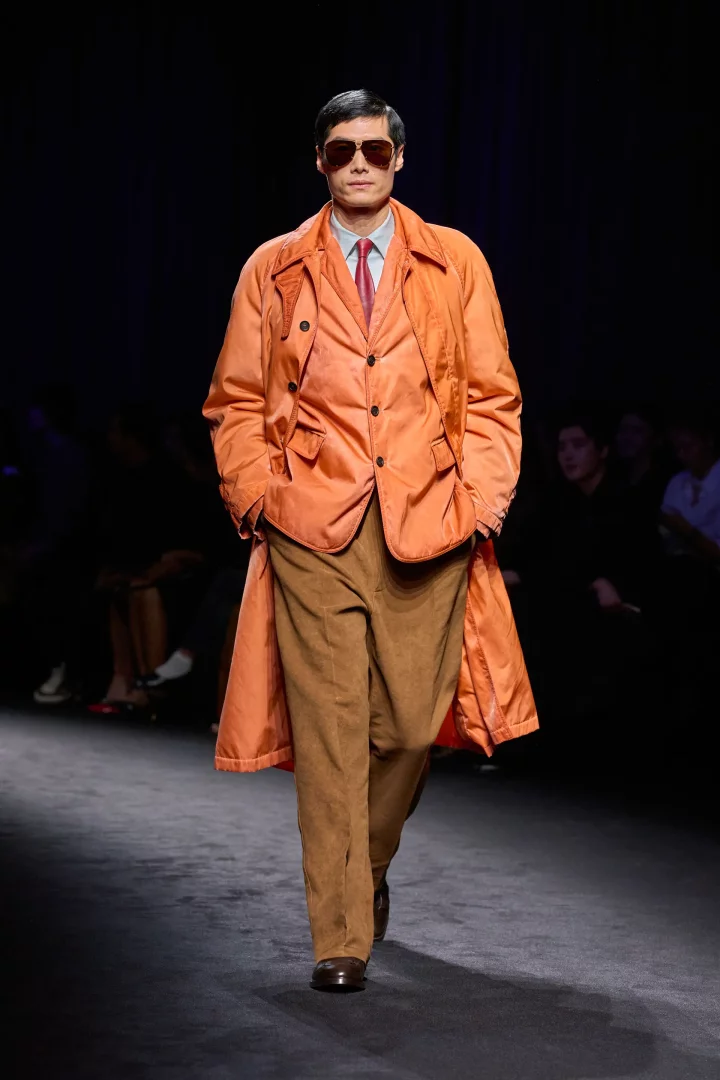 Ferragamo defilesinde 2026 sonbahar erkek modası renk trendlerinden alev turuncusu bir trençkot giyen model, koyu renk güneş gözlüğü, açık mavi gömlek, kırmızı kravat ve kahverengi bol pantolonuyla podyumda kendinden emin bir şekilde yürüyor.