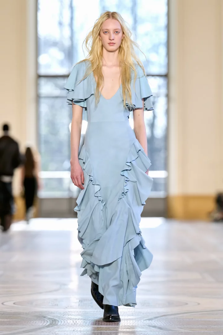 Uzun sarı saçlı bir model, Gabriela Hearst podyumunda fırfır detaylarıyla hareketlendirilmiş V yaka buz mavisi uzun bir elbise ve siyah botlarla yürüyerek Sonbahar Renk Trendleri arasında öne çıkan bu rengin zarif duruşunu sergiliyor.