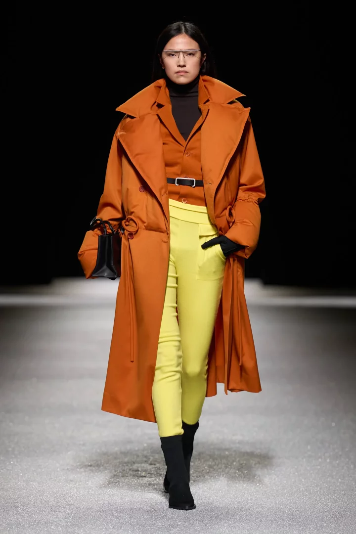 Podyumda yürüyen model, 2026 Sonbahar renk trendlerinden esintiler taşıyan Issey Miyake tasarımı geniş yakalı, kabarık turuncu bir palto (kadife çiçeği tonu) giyiyor. Siyah boğazlı kazağı ve turuncu gömleği tamamlayan parlak sarı dar paça pantolonu (şartröz), siyah botları ve küçük çantasıyla modern bir sonbahar kombini sergiliyor.