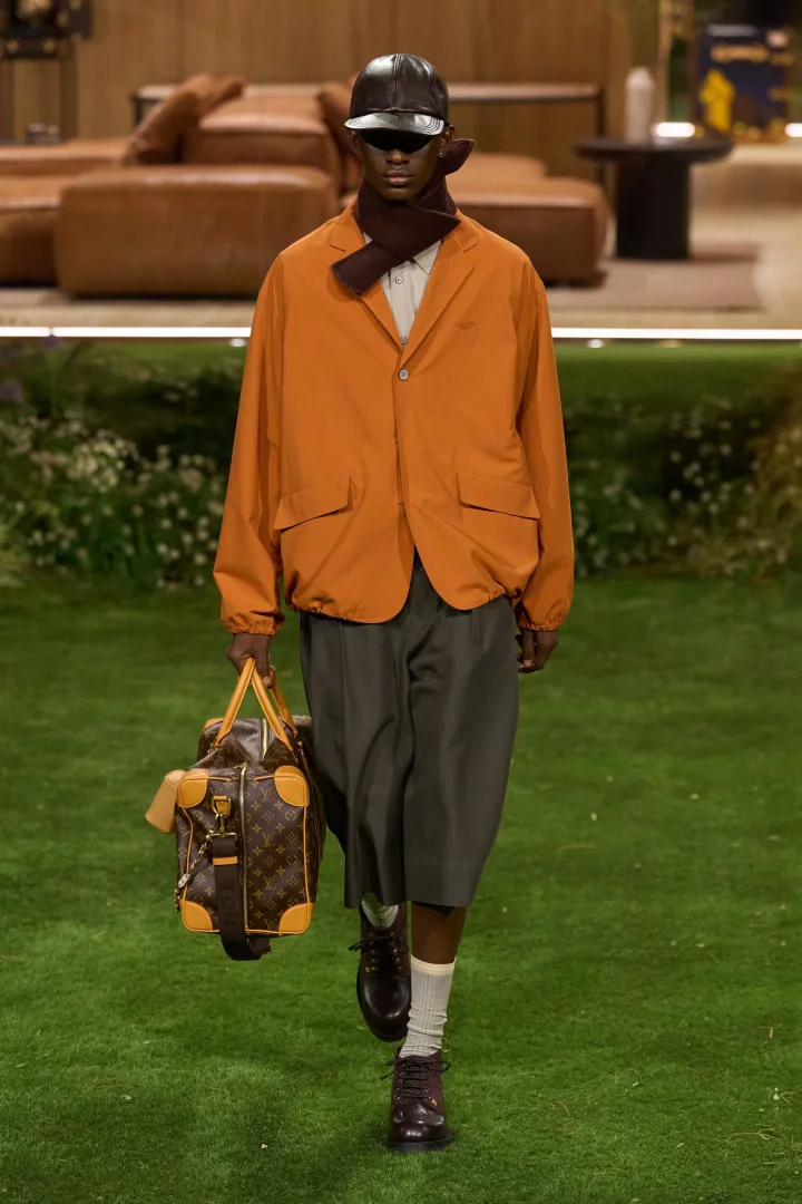 Bir Louis Vuitton defilesinde, 2026 sonbahar renk trendlerini yansıtan, alev turuncusu geniş kesim ceket, koyu füme şort, kahverengi şapka, eşarp, bej çoraplar ve bordo ayakkabılarla kombinlenmiş şık bir erkek modeli, desenli Louis Vuitton el çantasıyla yemyeşil bir podyumda yürüyor.
