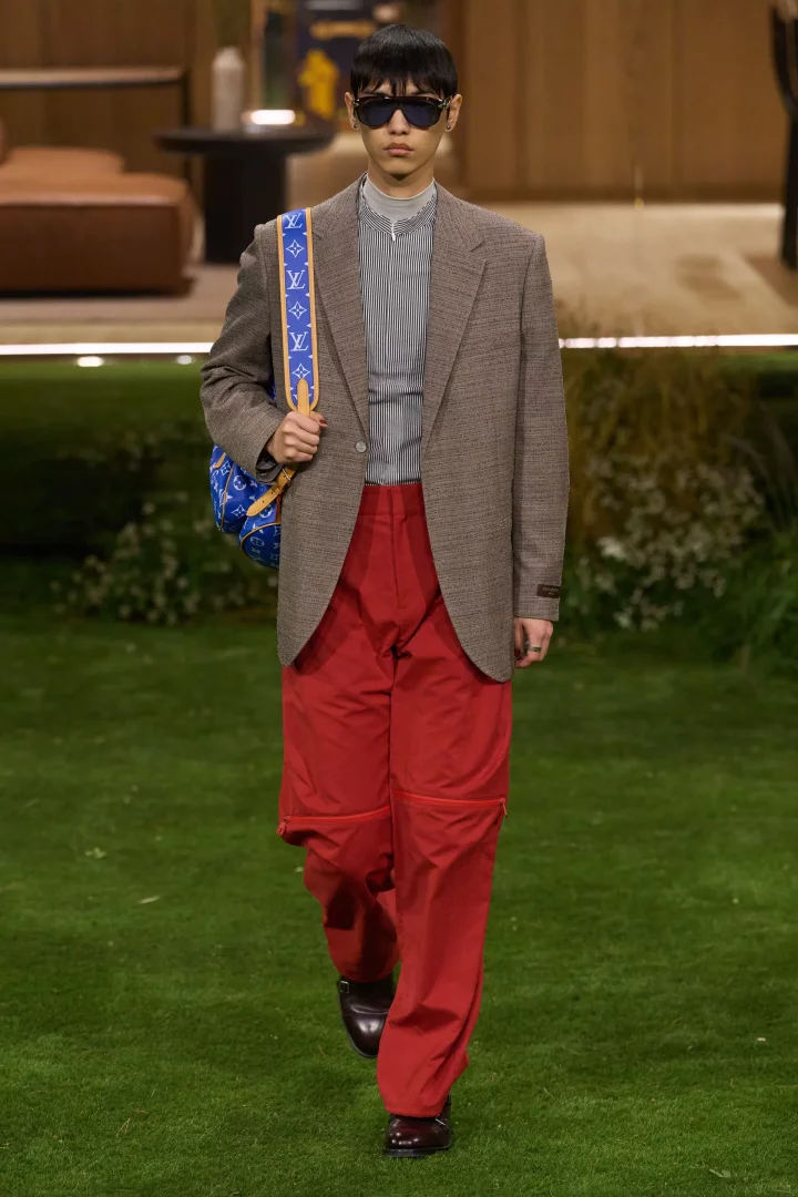 Louis Vuitton defilesinde, 2026 sonbahar renk trendlerini yansıtan bir erkek model, güçlü kırmızı bol pantolonu, toprak tonlarında blazer ceketi ve kobalt mavisi Louis Vuitton logolu çantasıyla podyumda yürüyor.