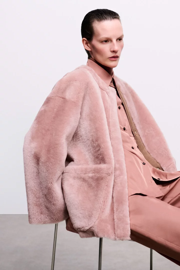 Model, geri taranmış saçları ve ciddi ifadesiyle, 2026 sonbahar renk trendlerinden biri olan toz pembe tonlarında, kürklü oversize bir ceket, saten bir gömlek ve pantolondan oluşan zarif bir kombinle beyaz arka plan önünde oturuyor.