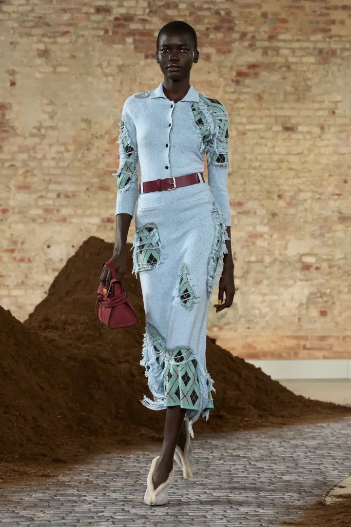 Koyu tenli bir model, 2026 sonbahar renk trendleri için Moschino podyumunda, buz mavisi rengi yıpranmış efektli, ekose desenli detaylara sahip, yakalı triko hırka ve midi etekten oluşan bir takım giyiyor, belinde bordo kemer, elinde bordo çanta ve tüylü, açık renkli terlik benzeri ayakkabılarıyla toprak yığınları ve tuğla duvar önünde yürüyor.