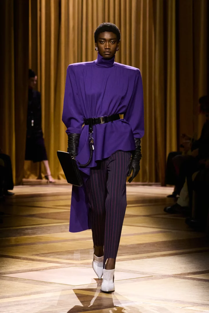 Mugler Sonbahar/Kış 2026-27 defilesinden, yüksek yakalı, omuzları vatkalı koyu mor bir bluz, çizgili pantolon, uzun siyah eldivenler ve beyaz botlarla podyumda yürüyen bir model, sezonun dikkat çeken koyu mor renk trendini vurguluyor.