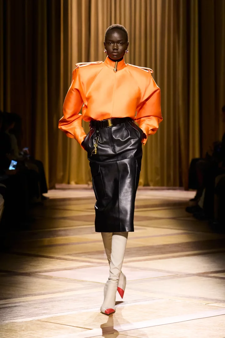 Mugler Sonbahar 2026 hazır giyim koleksiyonundan, podyumda yürüyen bir model, kabarık, vatkalı omuzlu parlak kayısı turuncusu üstü, altın tokalı siyah deri eteği ve kırmızı burunlu krem çizmeleriyle bu sonbaharın öne çıkan renk trendlerini gözler önüne seriyor.