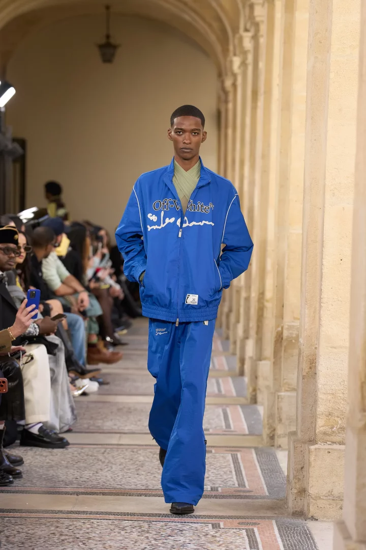 Bir podyumda yürüyen model, 2026 sonbahar renk trendlerinden biri olan Off-White markalı parlak kobalt mavisi bir eşofman takımı ve içinde şartröz benzeri açık yeşil bir üst giyerek sezonun cesur renk paletini sergiliyor.
