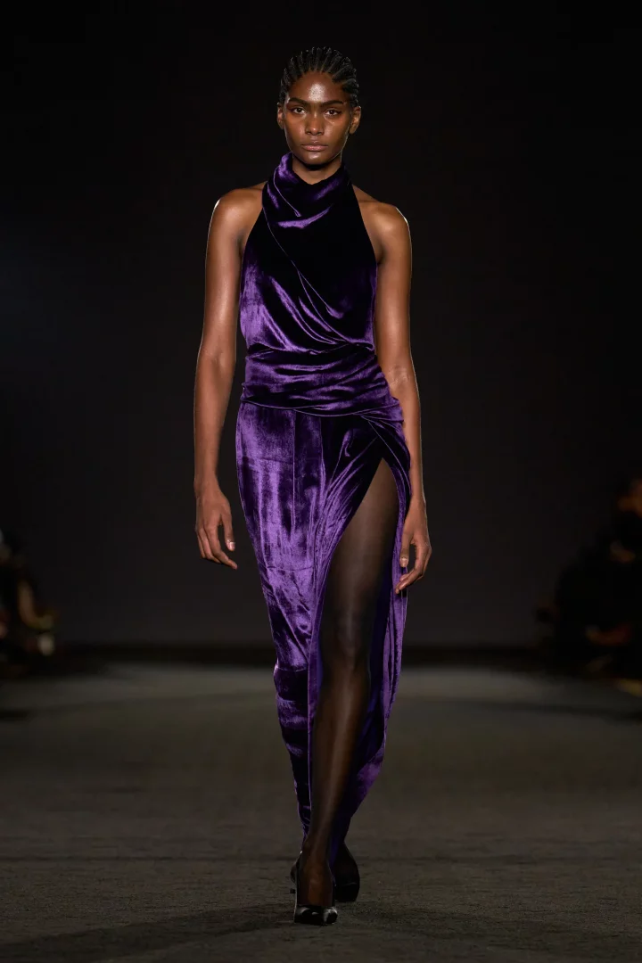 Koyu tenli bir model, Prabal Gurung defilesinde boyundan bağlamalı, drapeli ve derin yırtmaçlı koyu mor kadife elbisesiyle yürüyerek 2026 sonbahar renk trendlerinden biri olan morun gizemli duruşunu başarıyla yansıtıyor.