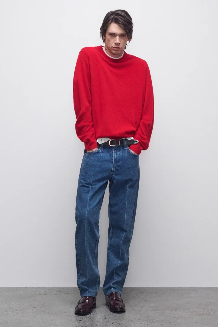 Rag & Bone 2026 Sonbahar Erkek Koleksiyonu'ndan, genç bir model parlak domates kırmızısı triko kazak, klasik mavi kot pantolon ve bordo makosen ayakkabılarla sade bir arka plan önünde durarak 2026 sonbahar renk trendlerinden canlı bir görünüm sergiliyor.