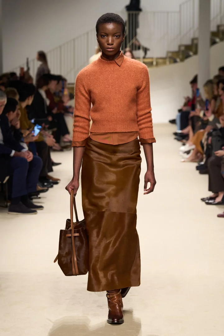 Bir model, Tod's Sonbahar 2026 Ready-to-Wear defilesinde podyumda yürüyor, sıcak kadife çiçeği renginde yün kazak ile parlak çikolata kahvesi tonlarında saten midi etek giyerek bu sezonun uyumlu sonbahar renk trendlerini sergiliyor, elinde kahverengi bir kova çanta ve kahverengi topuklu botlar giymiş.