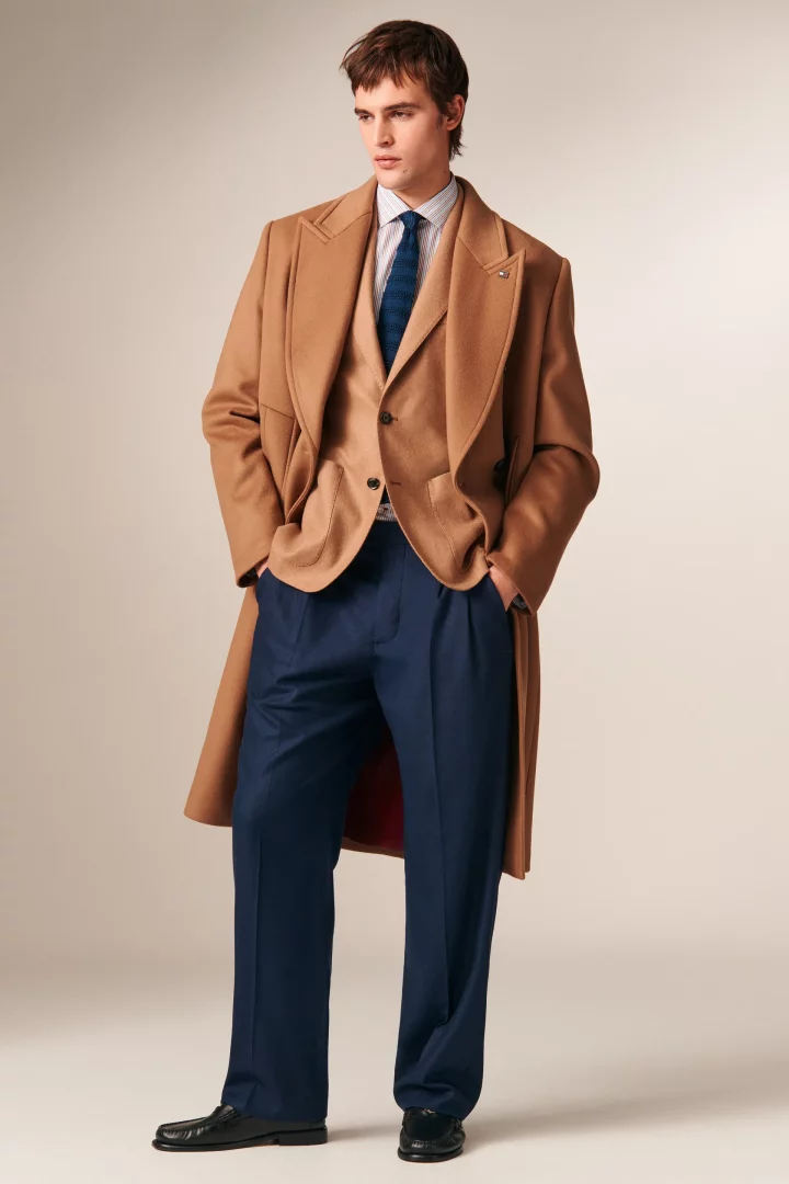 2026 sonbaharının renk trendlerini yansıtan bu şık erkek kombini, devetüyü bir kaban ve blazer ceket, açık mavi çizgili gömlek, kobalt mavisi desenli kravat ve lacivert pantolondan oluşuyor; genç adam elleri cebinde duruyor.