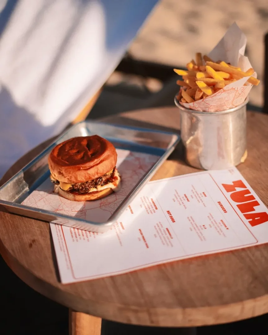 Yuvarlak ahşap bir masanın üzerinde, metal bir tepside kızarmış ekmekler arasında erimiş peynirli ve bol soslu iştah açıcı bir burger ile, yanında metal bir kapta servis edilmiş çıtır çıtır patates kızartmaları ve kırmızı Zula logolu bir menü kartı sıcak bir ortamda görülüyor.