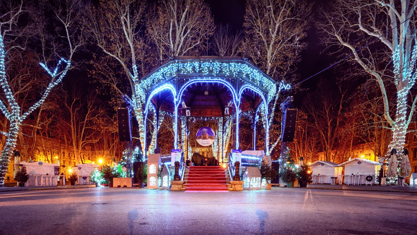 Zagreb'deki Advent kutlamaları sırasında gece çekilmiş bu geniş açılı fotoğrafta, mavi ve beyaz ışıklarla süslenmiş, kırmızı halılı bir çardak ve çevresindeki ışıklı ağaçlar ile Noel pazarı stantları görülüyor.