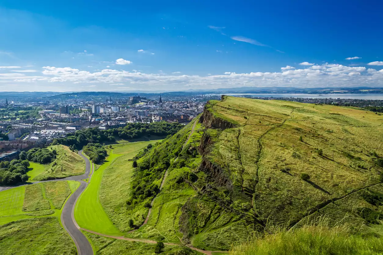 Mavi gökyüzü altında, Edinburgh şehrine bakan, gür yeşil otlarla kaplı sarp Arthurs Seat'ın yanı sıra, virajlı bir yolun geçtiği yemyeşil Holyrood Park'ı ve uzakta görünen denizi içeren panoramik bir manzara.