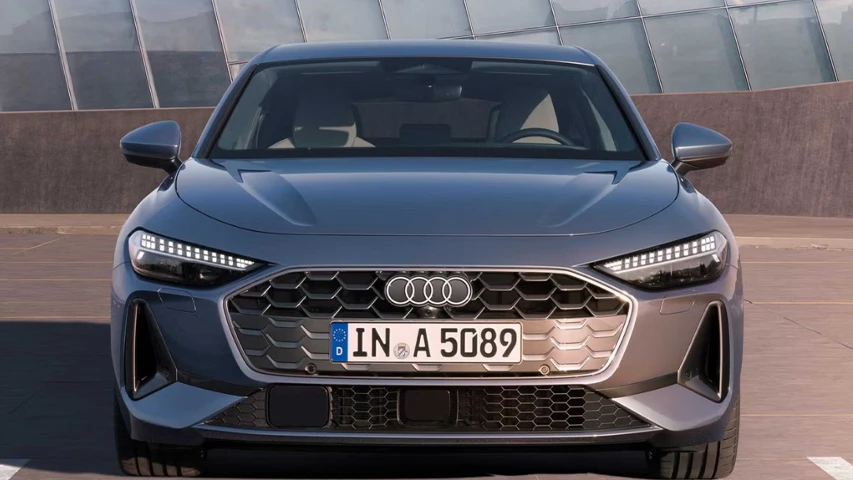 Koyu gri rengiyle dikkat çeken yeni nesil bir Audi A5 Sedan'ın ön cephesi, büyük altıgen ızgarası, belirgin LED farları ve ortasındaki dört halka logosuyla modern ve güçlü bir tasarım sergilerken, arka planda cam cepheli bir yapı duruyor.