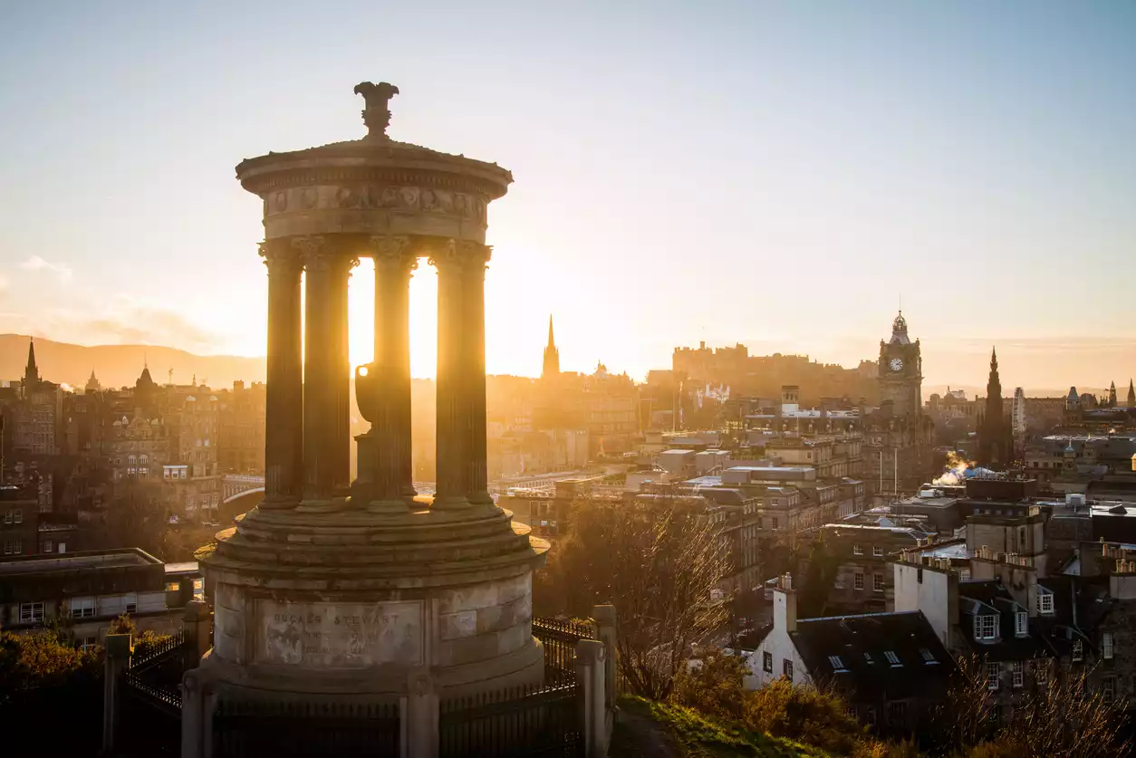 Calton Hill'den Edinburgh'un tarihi şehir manzarasını gösteren bu fotoğrafta, güneşin sütunlarının arasından parlayarak Dugald Stewart Anıtı'nı aydınlattığı, silueti belirgin kuleler ve Edinburgh Kalesi ile çevrili büyüleyici bir gün doğumu veya gün batımı anı yakalanmış.