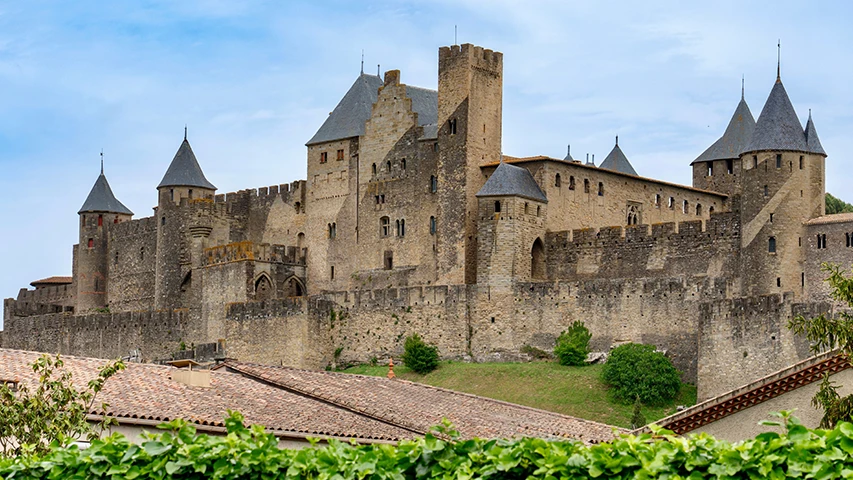 Mavi gökyüzü altında, eskimiş taş duvarları, burçları ve sivri çatılı sayısız kulesiyle heybetli Carcassonne Kalesi'nin, ön planda kiremit çatılı evler ve canlı yeşil bitki örtüsünün üzerinden yükselen tarihi ve etkileyici manzarası.
