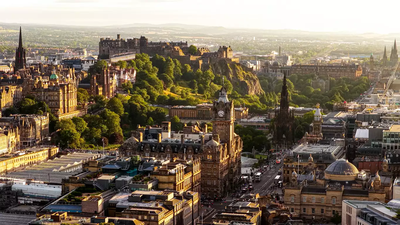 Edinburgh'un tarihi şehir merkezinin, gün batımı ışıkları altında parlayan Edinburgh Kalesi'nin heybetli duruşu, Scott Anıtı'nın sivri mimarisi ve Balmoral Otel'in saat kulesi gibi ikonik yapılarıyla dolu, yemyeşil ağaçlarla çevrili tepelerle süslenmiş panoramik bir kuşbakışı görünümü.