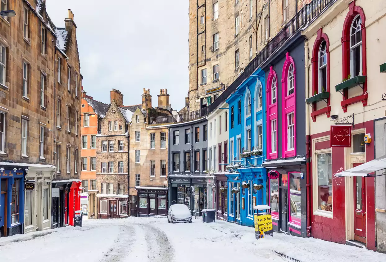 Edinburgh'un karlı bir sokağında, solda geleneksel taş binalar, sağda ise canlı mavi, pembe ve kırmızı renklere boyanmış dükkanlar sıralanıyor; yolda karla kaplı bir araç park etmiş, bu da şehrin kış atmosferini güzelce yansıtıyor.