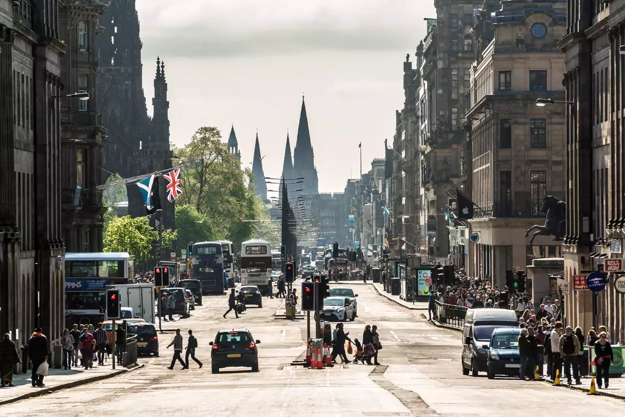 Edinburgh'un hareketli merkezindeki geniş bir caddenin genel görünümünde, Gotik mimariye sahip tarihi binalar, çift katlı otobüsler, araçlar ve karşıdan karşıya geçen yayalar dikkat çekerken, uzak planda Scott Anıtı ve diğer sivri kuleler şehrin siluetini belirginleştiriyor.
