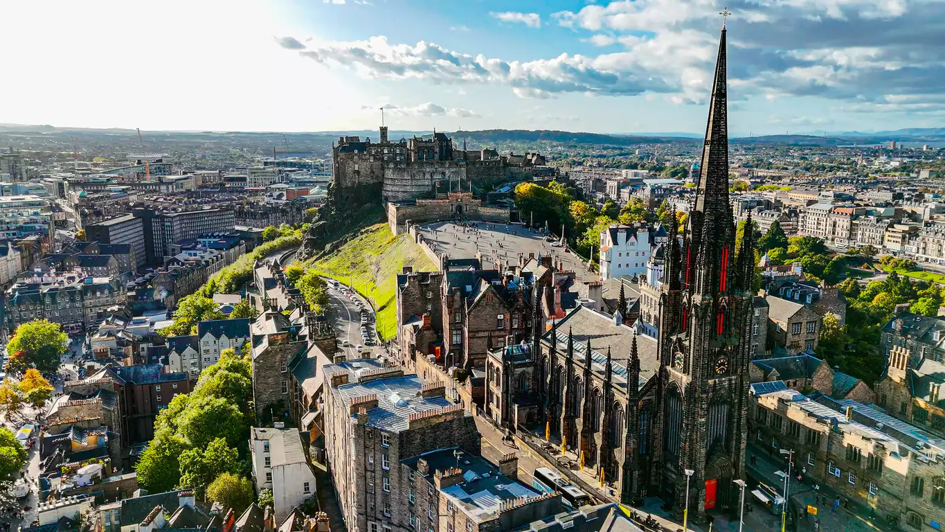 Güneşli bir günde, İskoçya'nın başkenti Edinburgh'un kuşbakışı görünümünde, şehrin simgesi olan kayalık bir tepe üzerindeki görkemli Edinburgh Kalesi ile sağda sivri gotik kulesiyle öne çıkan tarihi Tolbooth Kilisesi (The Hub), çevresindeki kalabalık meydanı ve yeşil alanlarla birlikte şehir manzarasına hakimdir.